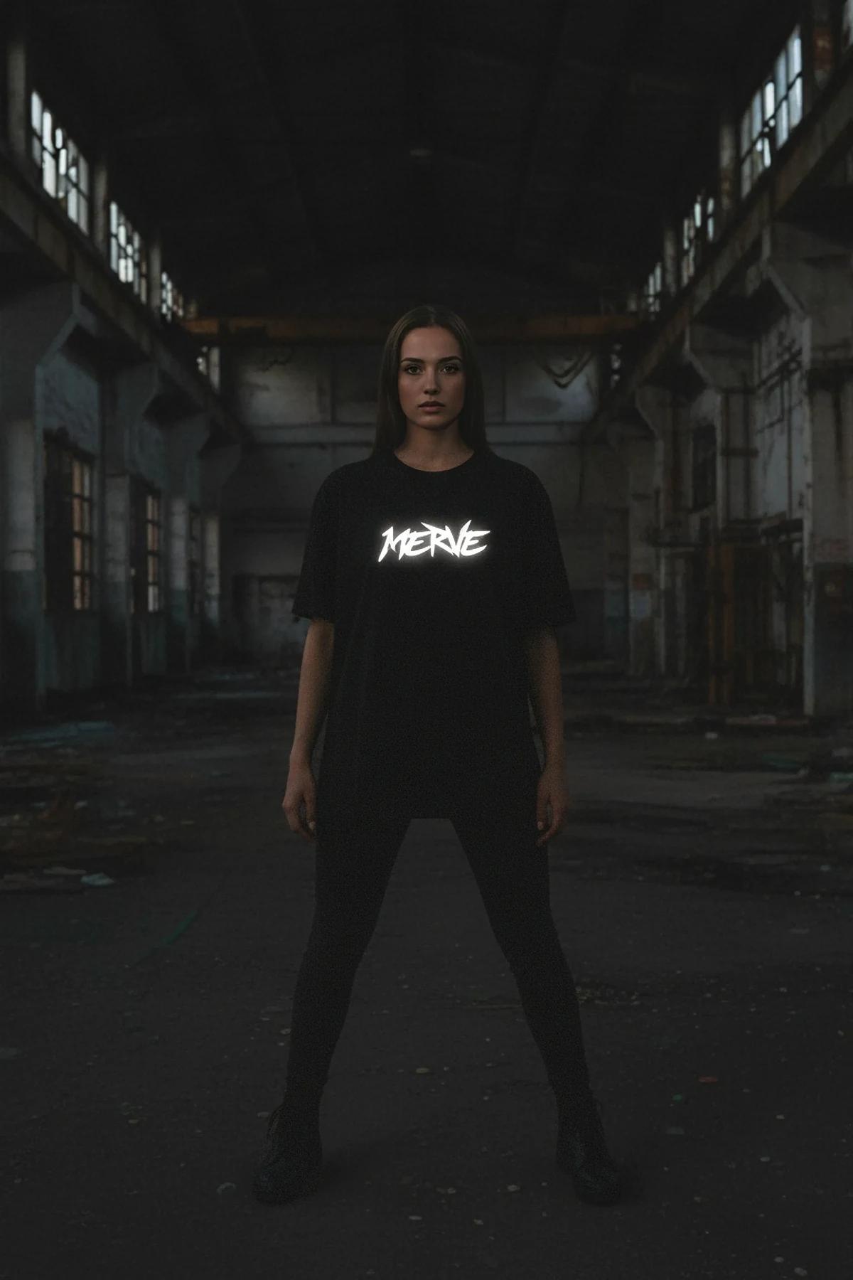 Oversized black t-shirt with a white reflective MERVE logo worn by a woman in an industrial setting.