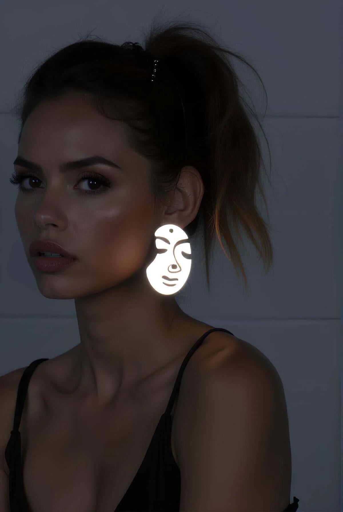 White reflective abstract face drop earrings glowing brightly under direct light worn with dark outfit
