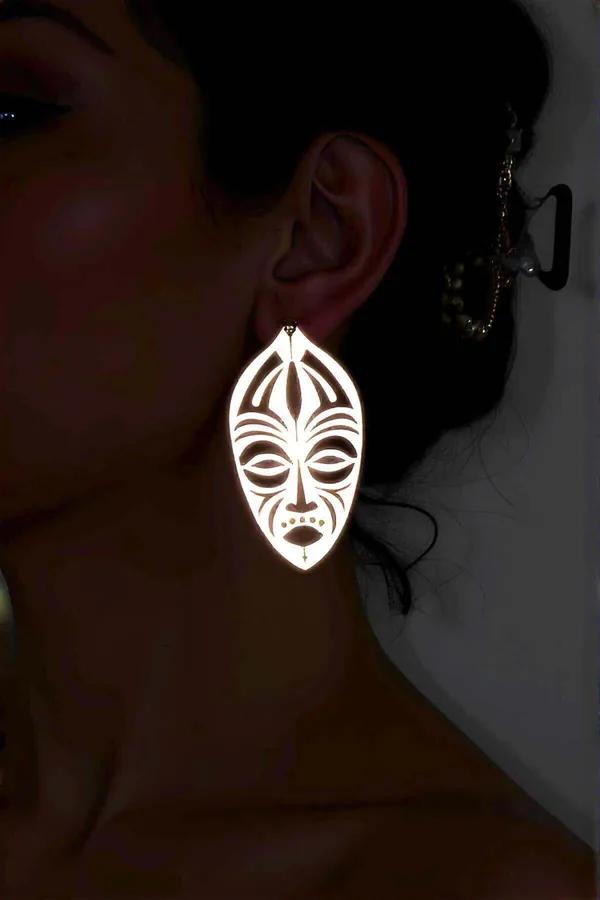 White reflective acrylic tribal mask drop earring with laser-cut face detail worn on ear against dark background
